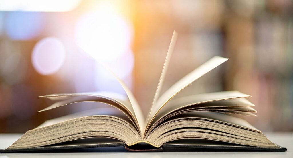 An open book with its pages fanned out sits on a table. The background is softly blurred with warm, glowing light and circular bokeh, creating a cozy and inviting atmosphere.