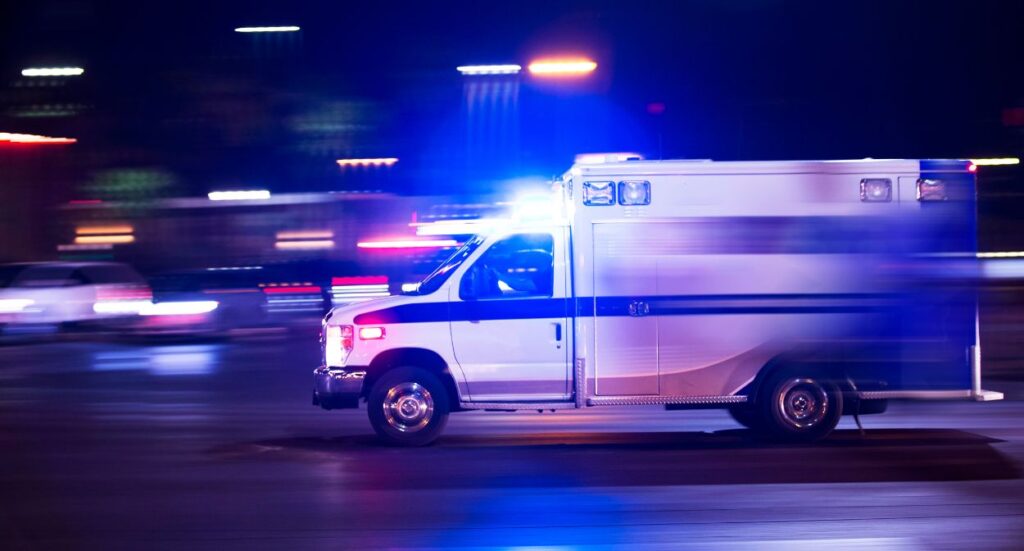 An ambulance with flashing lights speeds down a city street at night, with blurred lights and cars in the background indicating motion and urgency.