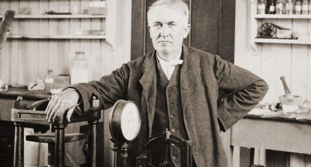 A man in a suit stands beside scientific instruments in an old laboratory, with shelves of bottles and equipment visible in the background.