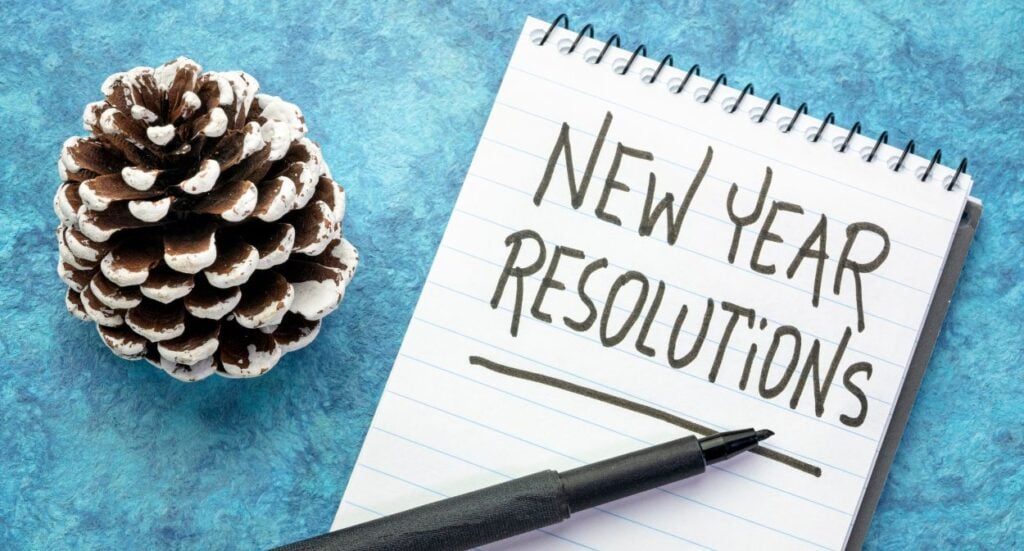 A black pen rests on a notepad with “New Year Resolutions” written on it in bold letters. A frosted pinecone is placed to the left, all set on a textured blue surface.