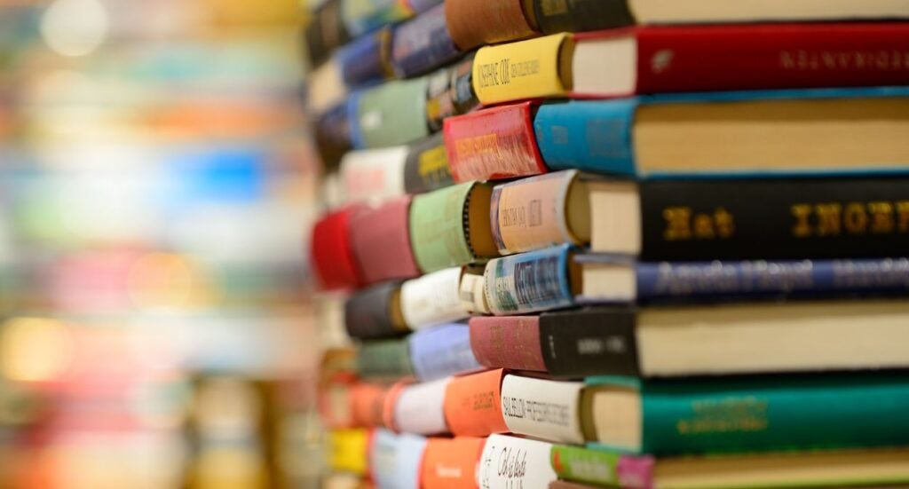 A close-up of several stacks of books piled on top of each other, with colorful covers and spines. The background is blurred, highlighting the variety of books in the foreground.