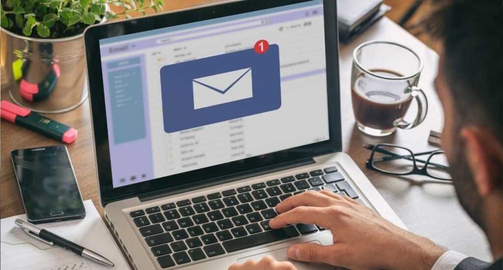 A person checks their email on a laptop at a desk, with a large email notification icon on the screen. The desk has a coffee cup, notebook, pen, glasses, and a plant.