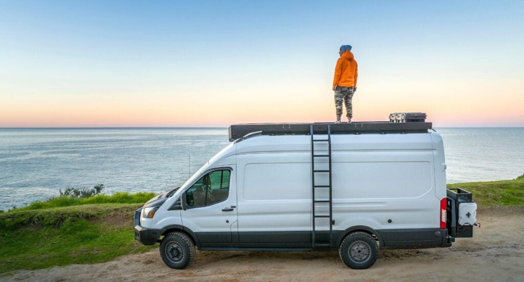 A person in an orange jacket stands on top of a white camper van parked near a grassy cliff, overlooking the ocean at sunset. A ladder is attached to the van, and the sky is clear and pastel-colored.