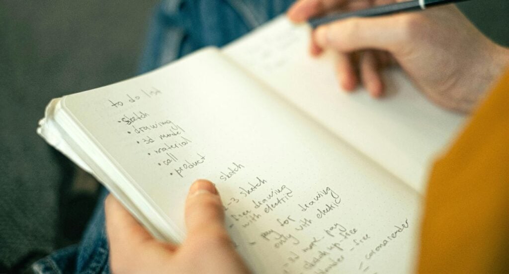 A person writes in a notebook, holding a pen over a handwritten to-do list with tasks like “sketch,” “drawing,” “read manual,” and “inspiration.” The notebook is open and held on their lap.