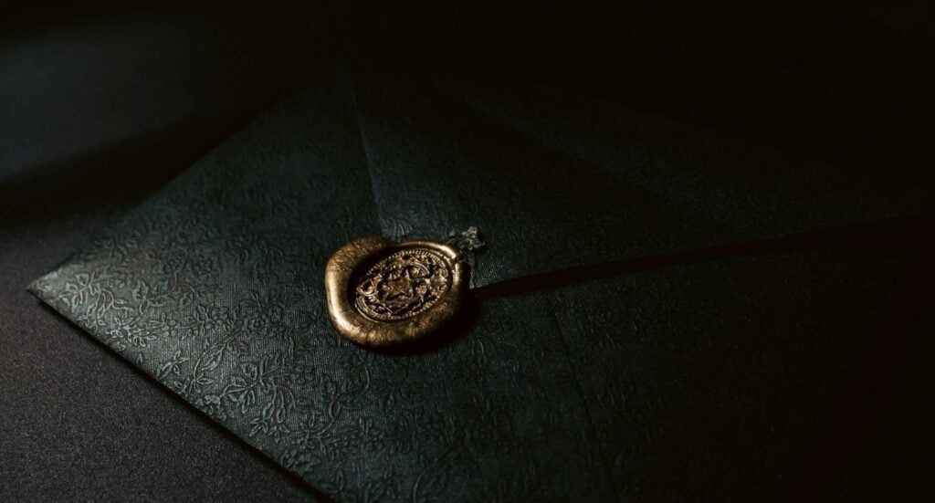 A dark, textured envelope sealed with an ornate gold wax seal, resting on a dark surface with dramatic lighting highlighting the seal and envelope pattern.