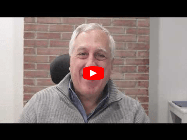 A person with gray hair smiles while sitting in front of a brick wall, wearing a gray sweater. A red play button is centered on their face, indicating a video thumbnail.
