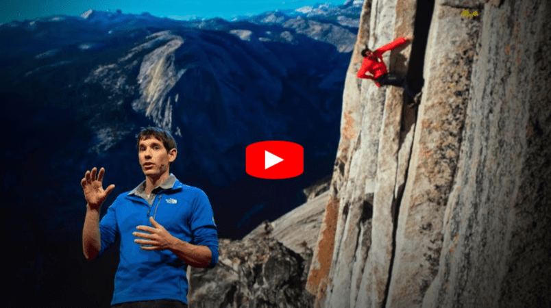 A man in a blue jacket speaks on stage, with an image of a rock climber scaling a steep cliff and a mountainous landscape in the background. A red play button icon appears in the center, indicating a video.