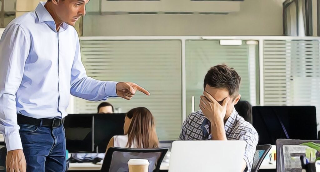 A man standing and pointing angrily at a seated coworker, who covers his face in frustration at his desk in a modern office. Other employees work in the background.