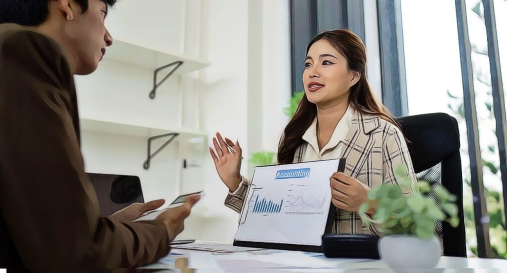 A woman in a plaid blazer shows an accounting chart with graphs to a man during a business meeting in a bright office with plants and shelves in the background.