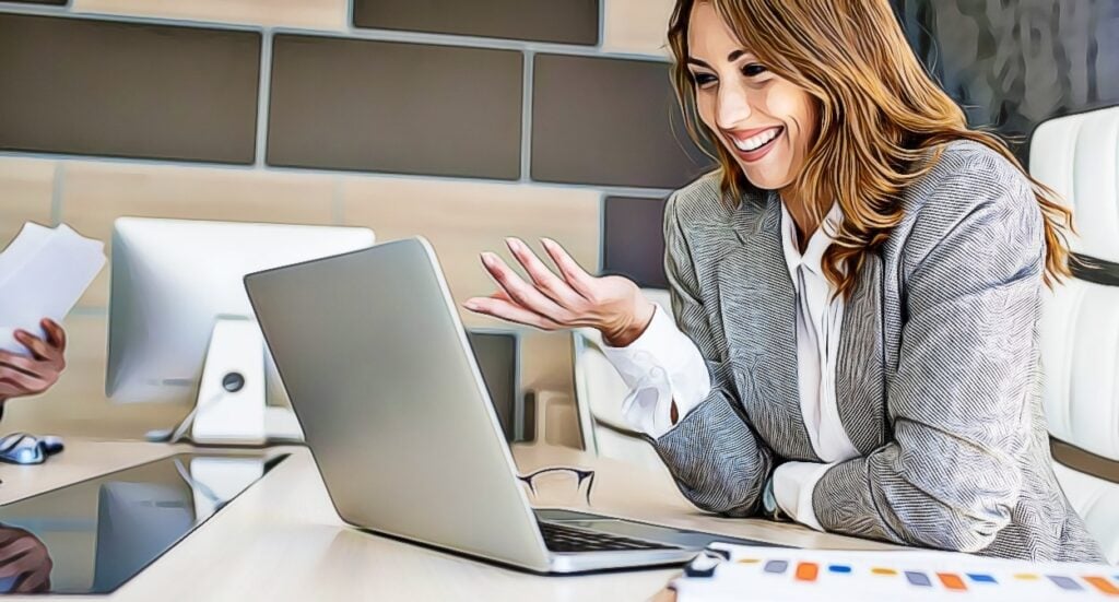A woman in a gray blazer smiles and gestures while sitting at a desk with a laptop, having a virtual meeting in a modern office.
