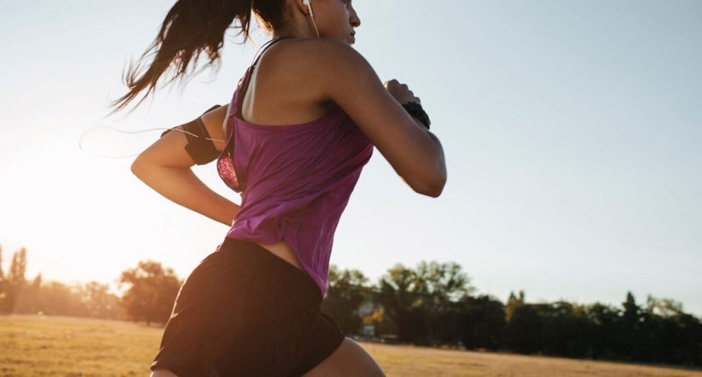 A woman in athletic clothing runs outdoors on a grassy field during sunset, wearing earphones and an armband, with trees and a clear sky in the background.
