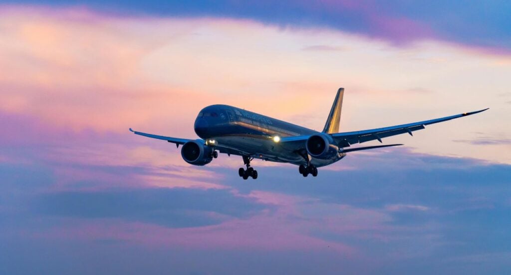 A commercial airplane is flying in the sky at sunset, with its landing gear down and lights on. The sky is filled with pink, purple, and blue clouds.