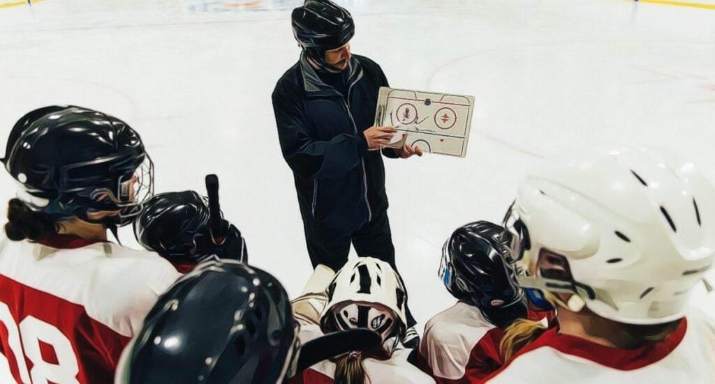 A hockey coach in black gear holds a clipboard and explains strategy to a group of players in white and red uniforms gathered around him on the ice rink.