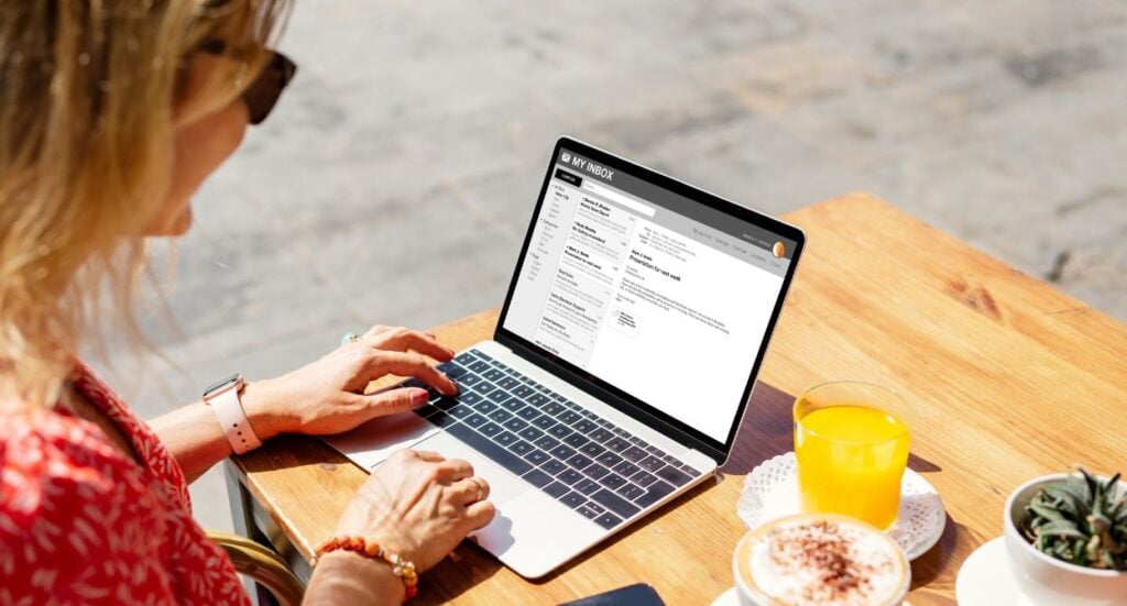 A woman sits at a wooden table outdoors, using a laptop with an email inbox open. On the table are a glass of orange juice, a cup of coffee with foam, and a small potted plant.