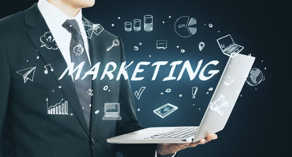A person in a suit holds a laptop with the word MARKETING and various marketing-related icons, such as graphs, a megaphone, and charts, sketched around the text on a dark background.