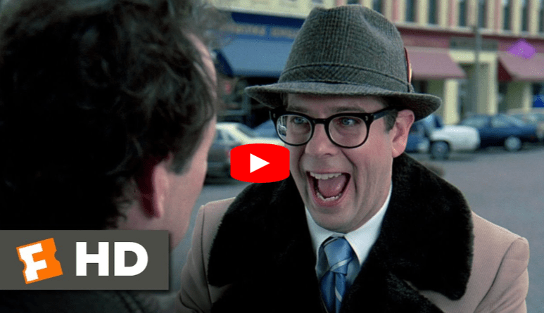 A man in a hat, glasses, and overcoat excitedly smiles at another person on a city street; HD and Fandango logos appear in the lower left, with a YouTube play button overlay in the center.