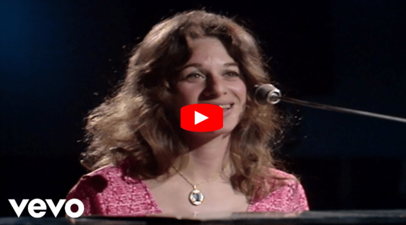 A woman with wavy brown hair, wearing a pink top and a cameo necklace, smiles while sitting at a piano and singing into a microphone. The Vevo logo is visible in the lower left corner.