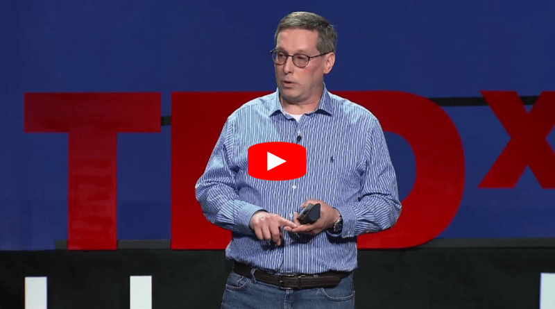 A man in glasses and a striped blue shirt speaks on stage in front of a large red TEDx sign, holding a device in his hand.