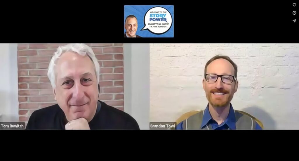 Two men smiling during a video call. The man on the left has short white hair and a black shirt, sitting in front of a brick wall. The man on the right has short brown hair, glasses, and a collared shirt, sitting in front of a white wall.