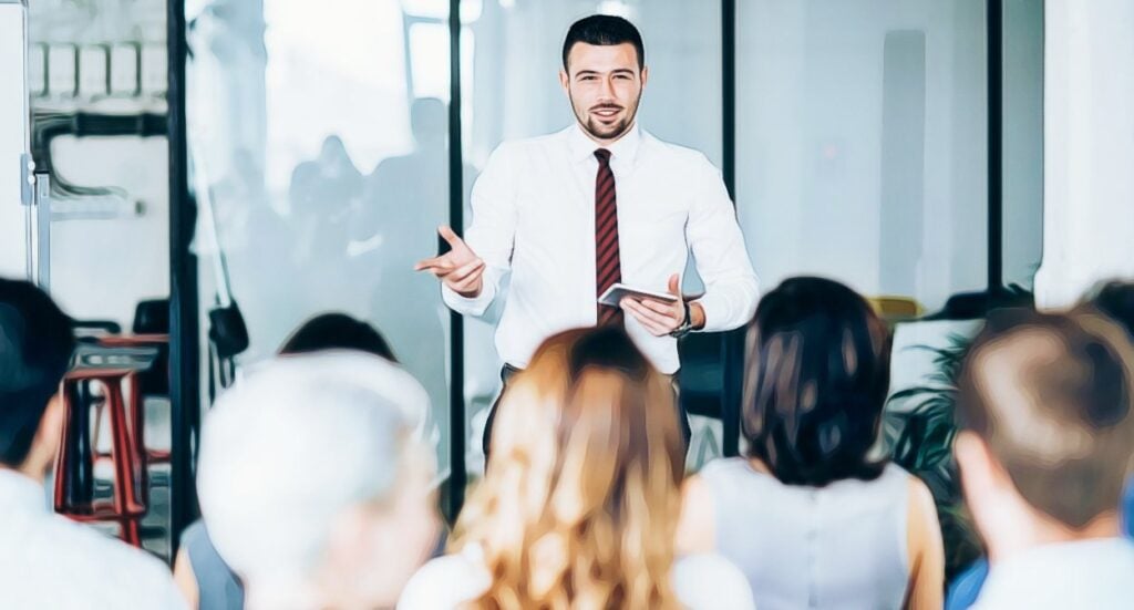 A man in a white shirt and red tie stands at the front of a room, speaking to an audience seated and listening attentively. He holds a tablet and gestures with his hand. The setting appears to be an office or conference room.